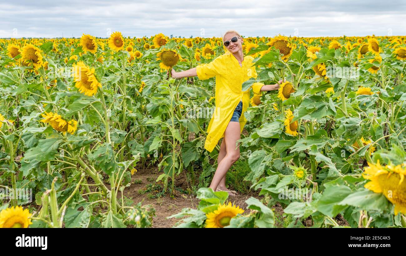 Happy lady in a yellow dress adult with a lovely smile on the background of sunflowers blue clouds in hot sunny weather Stock Photo
