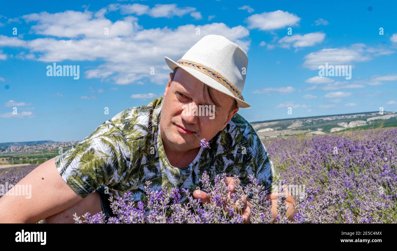 Smiling guy in the lilac field looks seriously at the flowers sitting in a cap against the blue sky in the afternoon Stock Photo