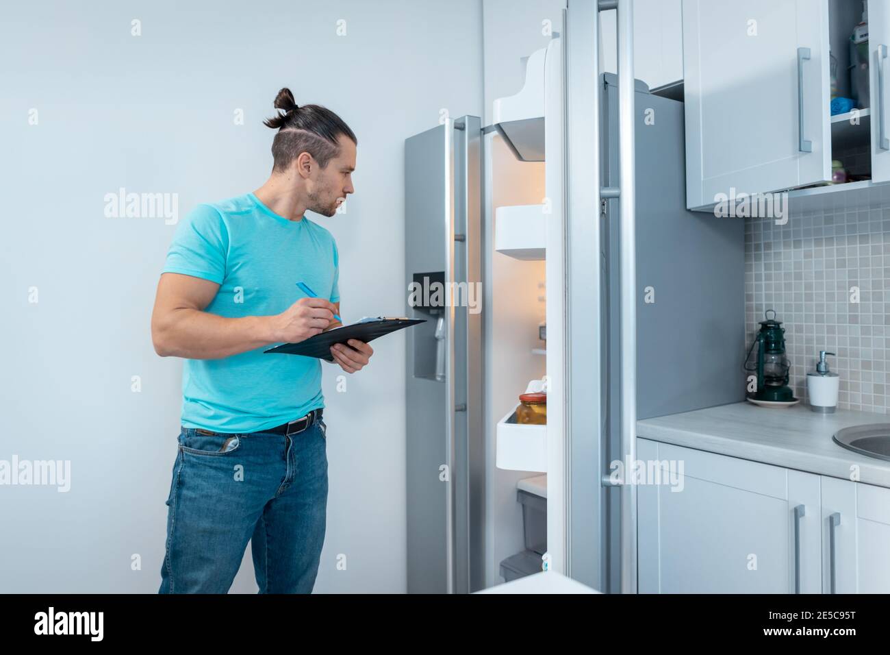 fitness nutritionist man with clipboard revising or look at fridge at kitchen Stock Photo