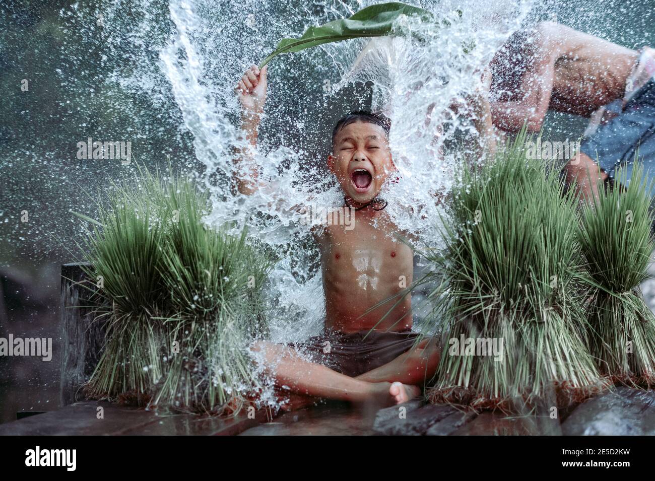 Boy sitting on a jetty being splashed with water by his father, Thailand Stock Photo