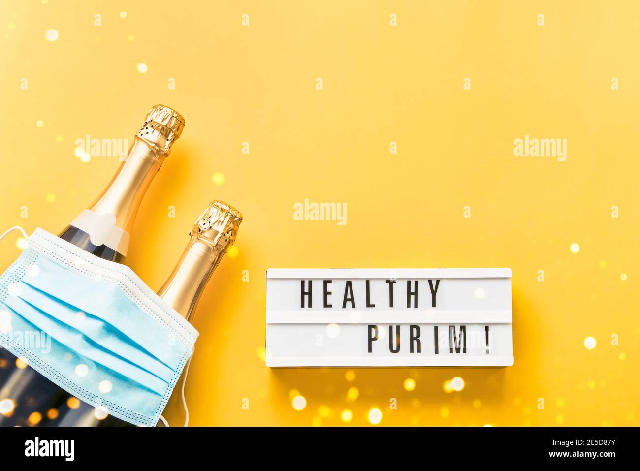Healthy Purim written in lightbox, two champagne bottles, and medical mask on a yellow background. Stock Photo