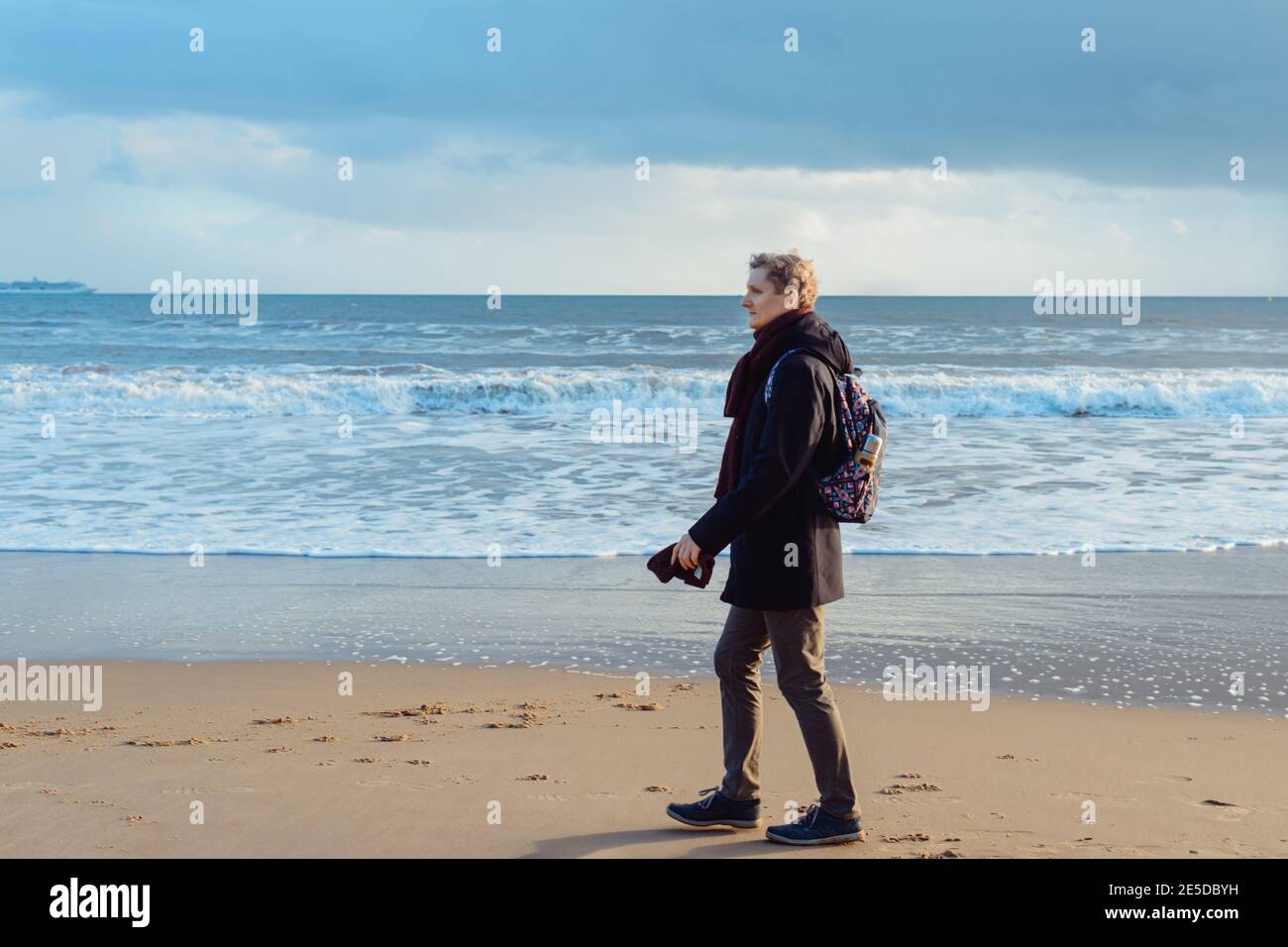 A young man in warm clothes with backpack walking on winter seaside and enjoying the moment. Relax during a walk on the coast. Simple pleasures Stock Photo