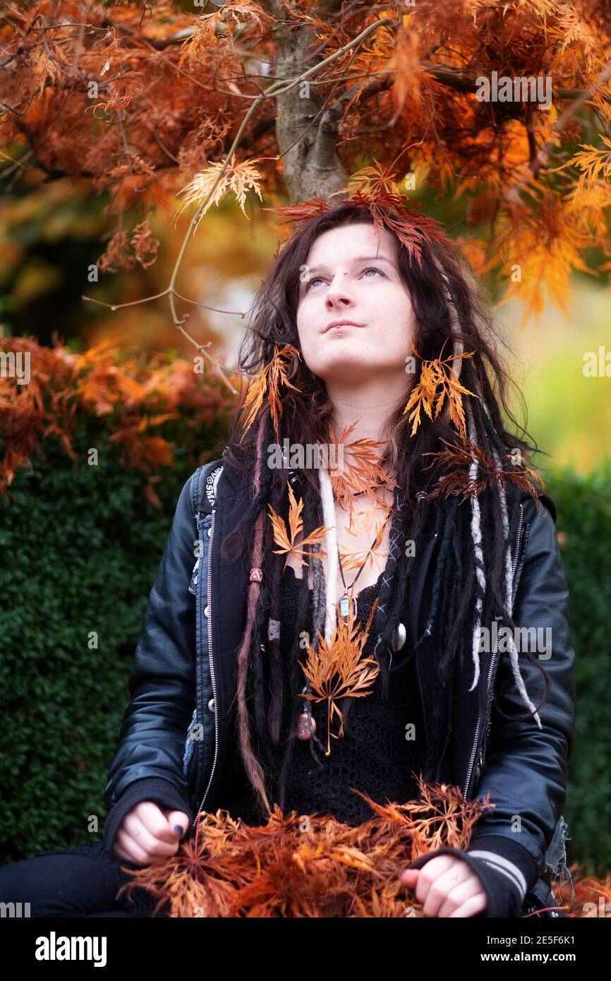 Beautiful young individual, eccentric woman, with dreadlocks, piercing and tattoo, with black leather jacket sits under the Japanese maple tree and is Stock Photo