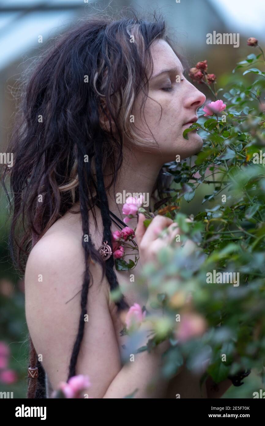 Beautiful young individual, eccentric woman, with interesting hairstyle, dreadlocks, piercing and tattoo smelling in garden park with closed eyes of p Stock Photo