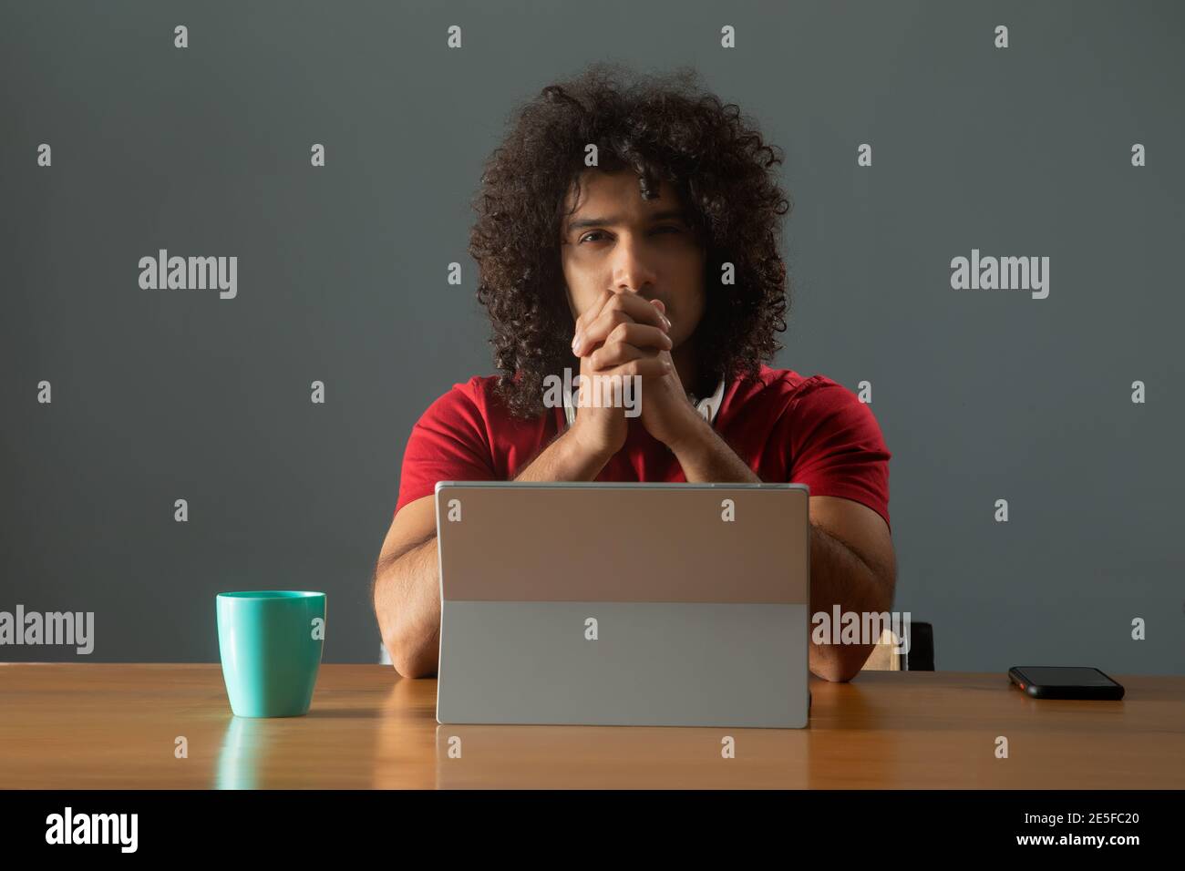 A CURLY HAIRED YOUNG MAN LOOKING AT CAMERA WHILE SITTING IN FRONT OF DIGITAL TABLET Stock Photo