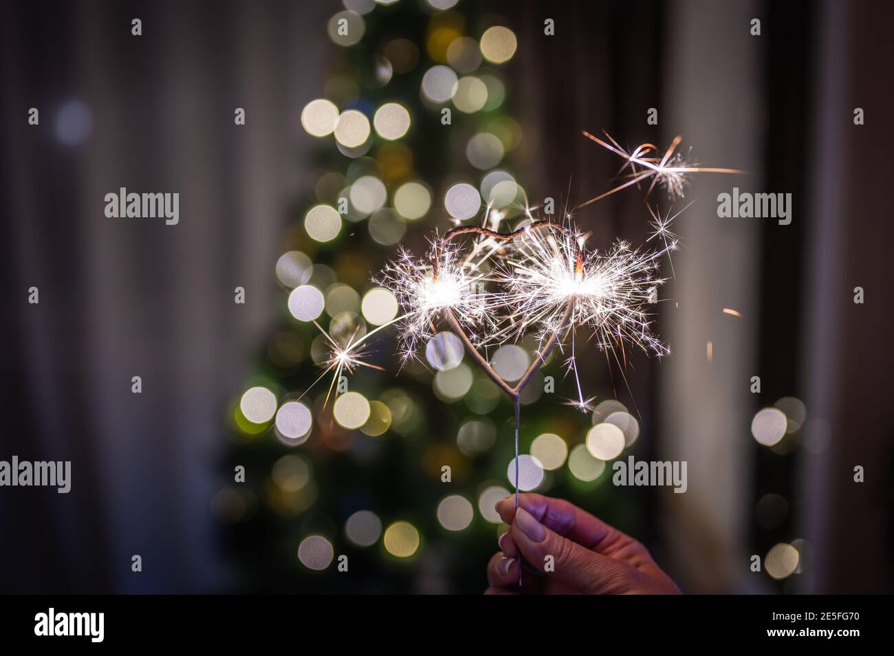 Bengal fire heart-shaped in the woman's hand burns against the background of a Christmas tree with colored garlands with bokeh Stock Photo