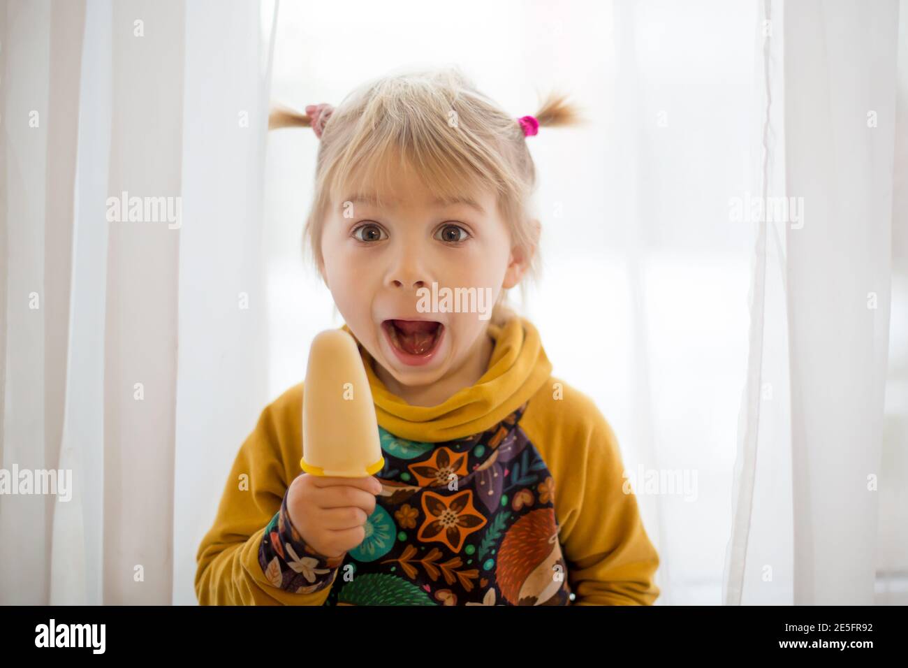 Cute sweet child, toddler with little ponytails, goofying at home, eating ice cream, making funny faces and smiling Stock Photo