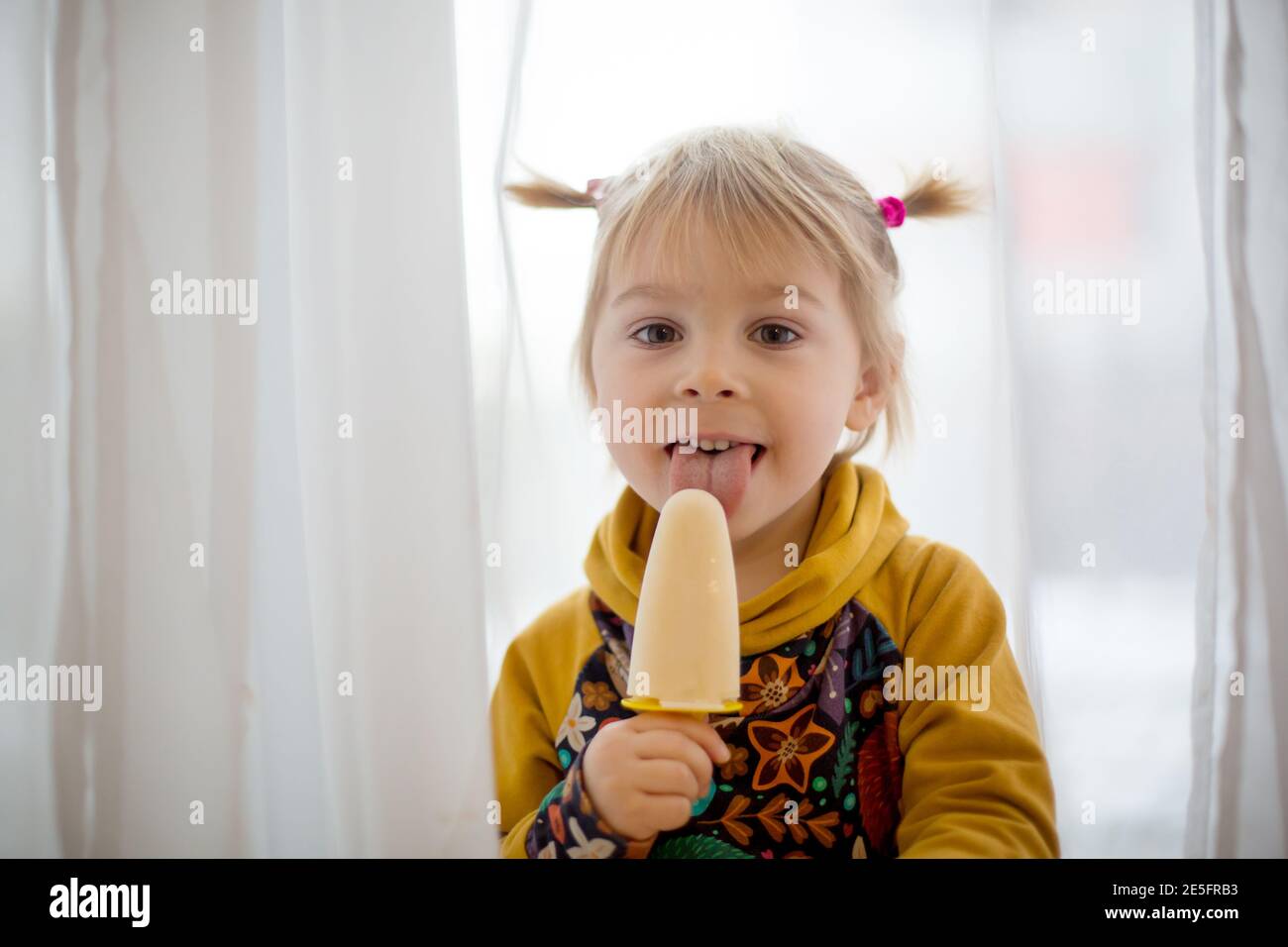 Cute sweet child, toddler with little ponytails, goofying at home, eating ice cream, making funny faces and smiling Stock Photo