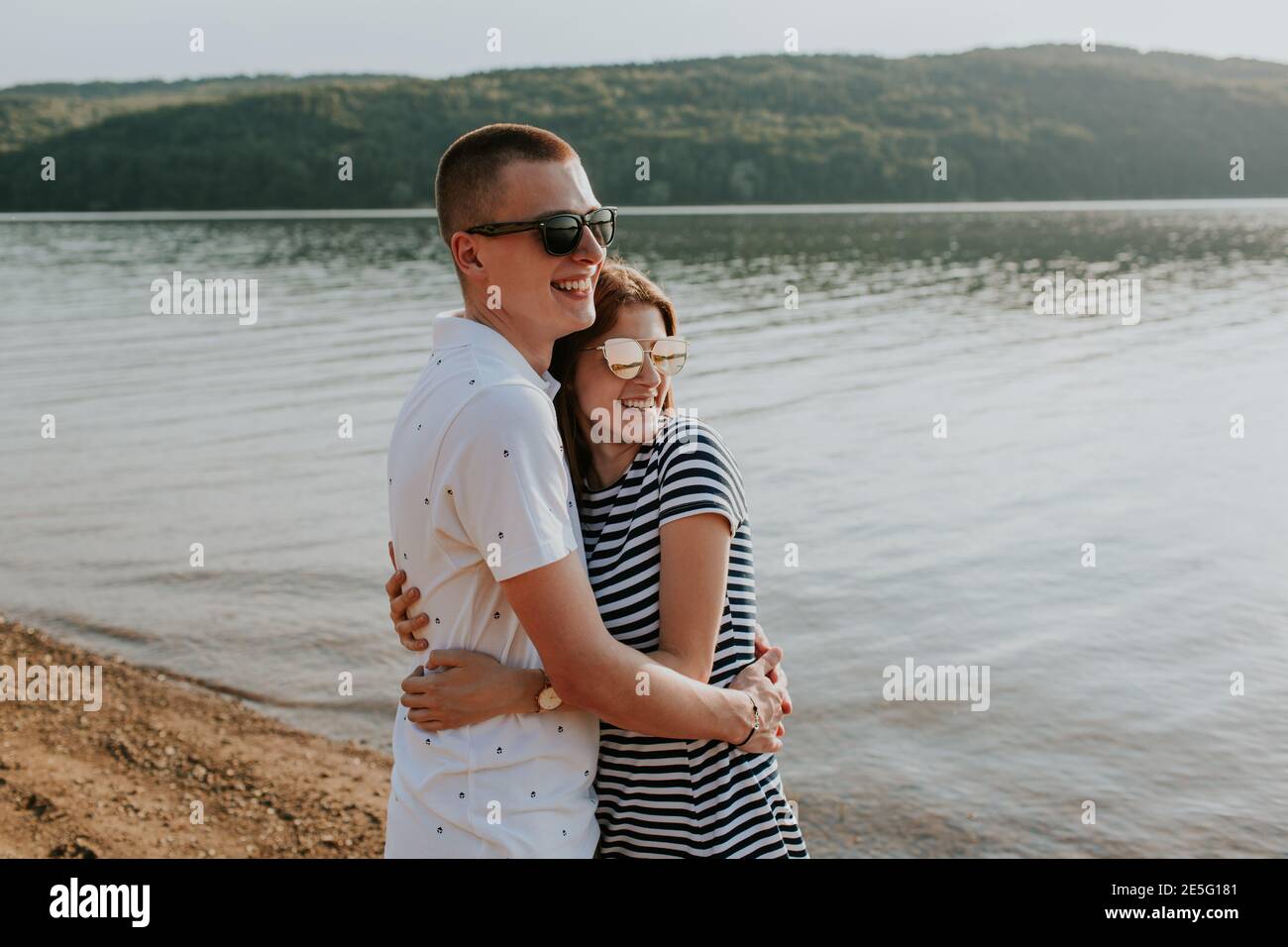 Cheerful couple in love hugging at the lake at sunset. Portrait of smiling boyfriend and girlfriend embracing on the beach on warm summer evening. Stock Photo