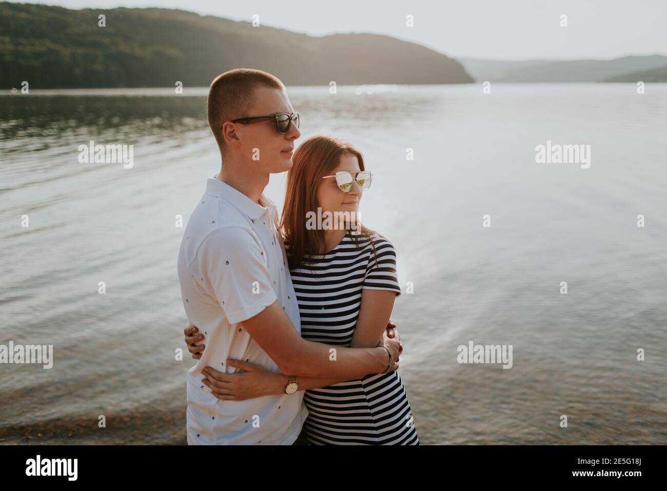 Couple in love hugging at the lake at sunset. Portrait of smiling boyfriend and girlfriend embracing on the beach on warm summer evening. Stock Photo