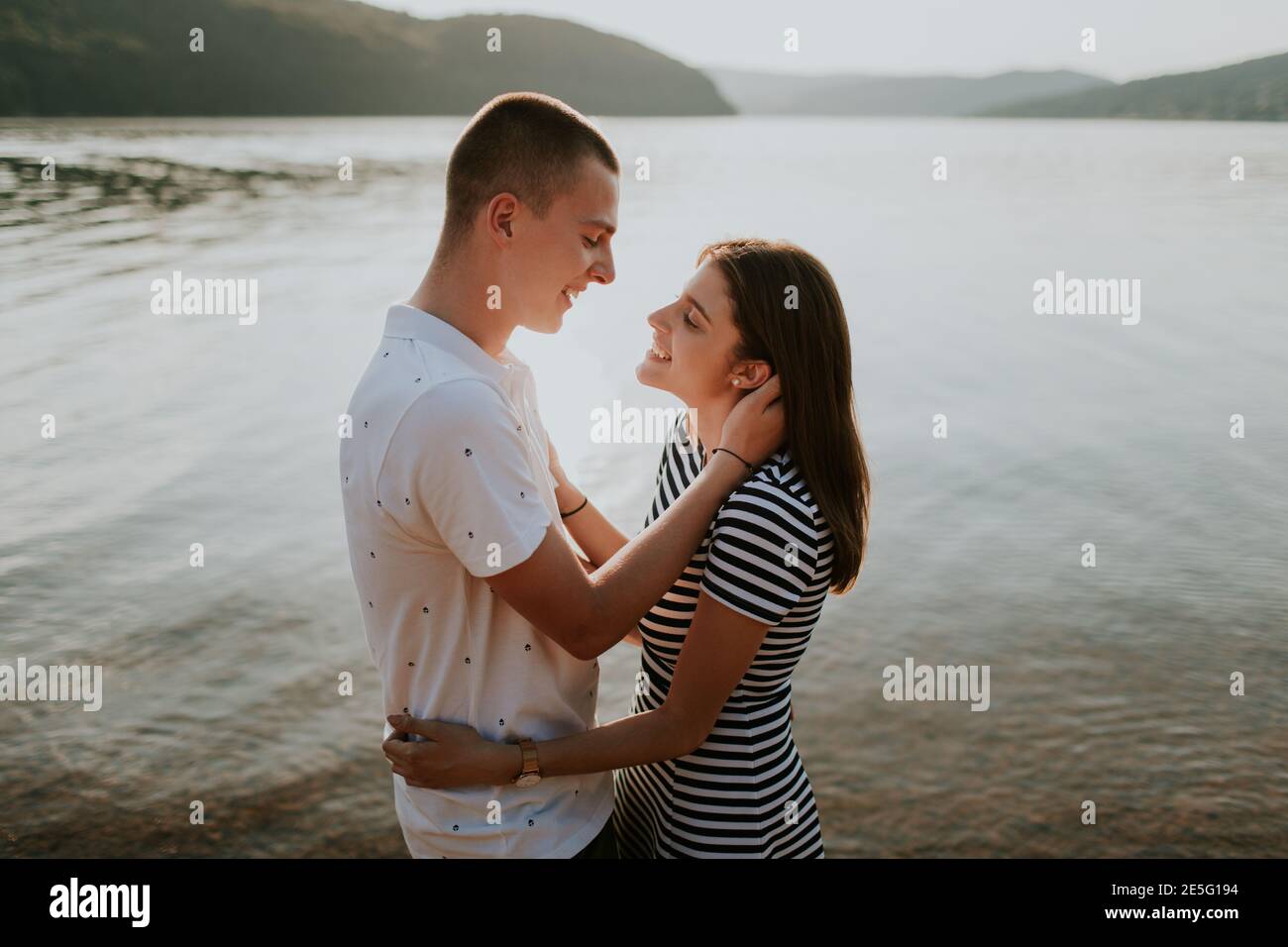 Cheerful couple in love hugging at the lake at sunset. Portrait of smiling boyfriend and girlfriend about to kiss on the beach on warm summer evening. Stock Photo
