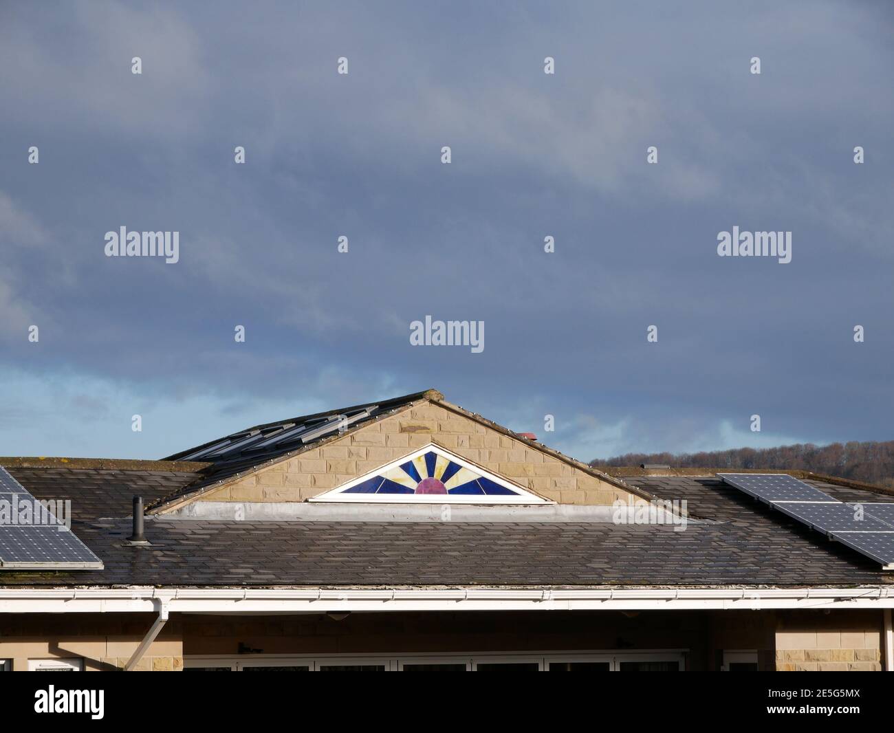 House roof with sun burst triangular stained glass window in blue yellow and red with a blue sky above Stock Photo