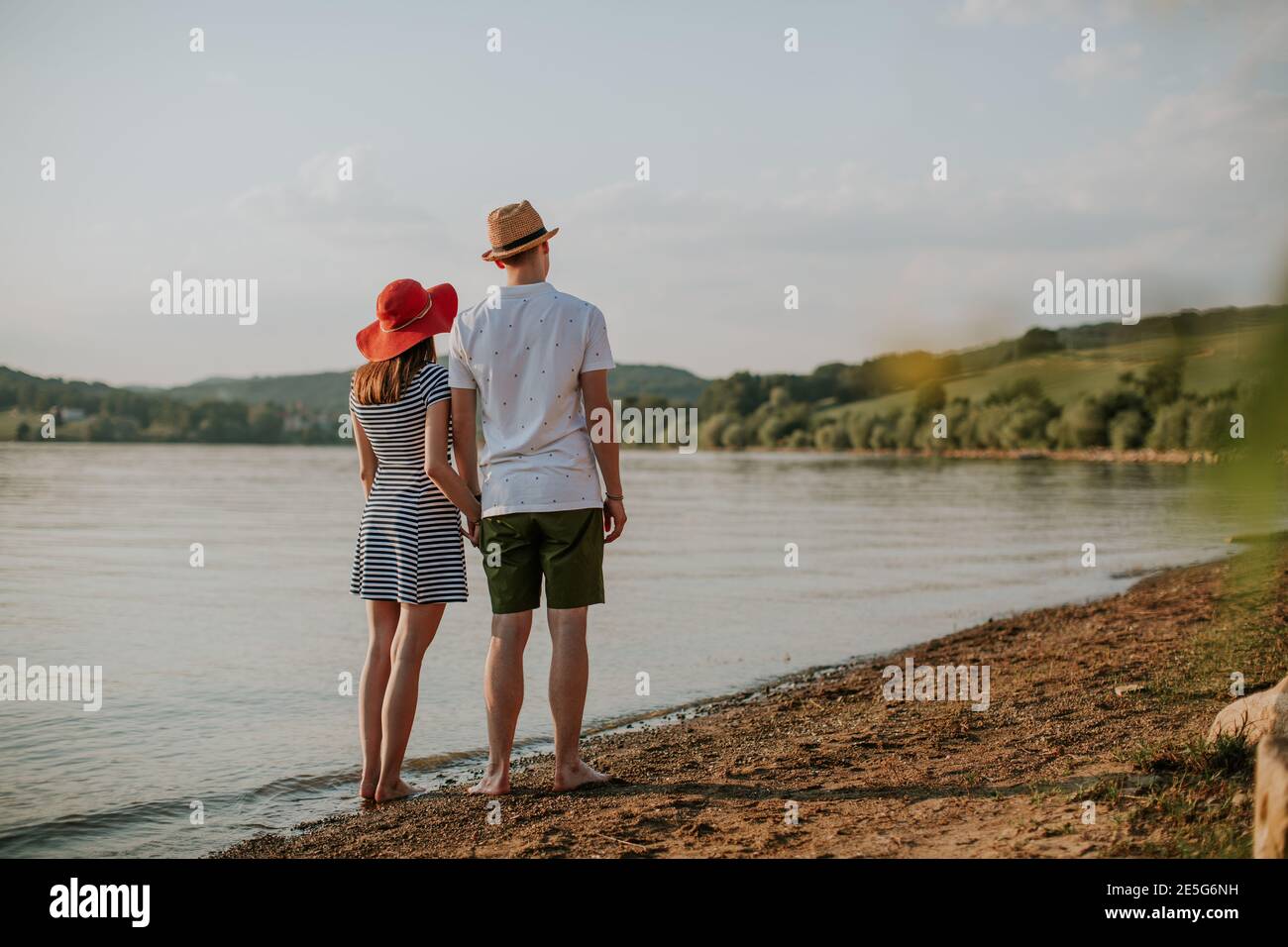 Back view of couple holding hands and going for walk on the beach at sunset. Rear view of man and woman standing at the lake on summer evening. Stock Photo