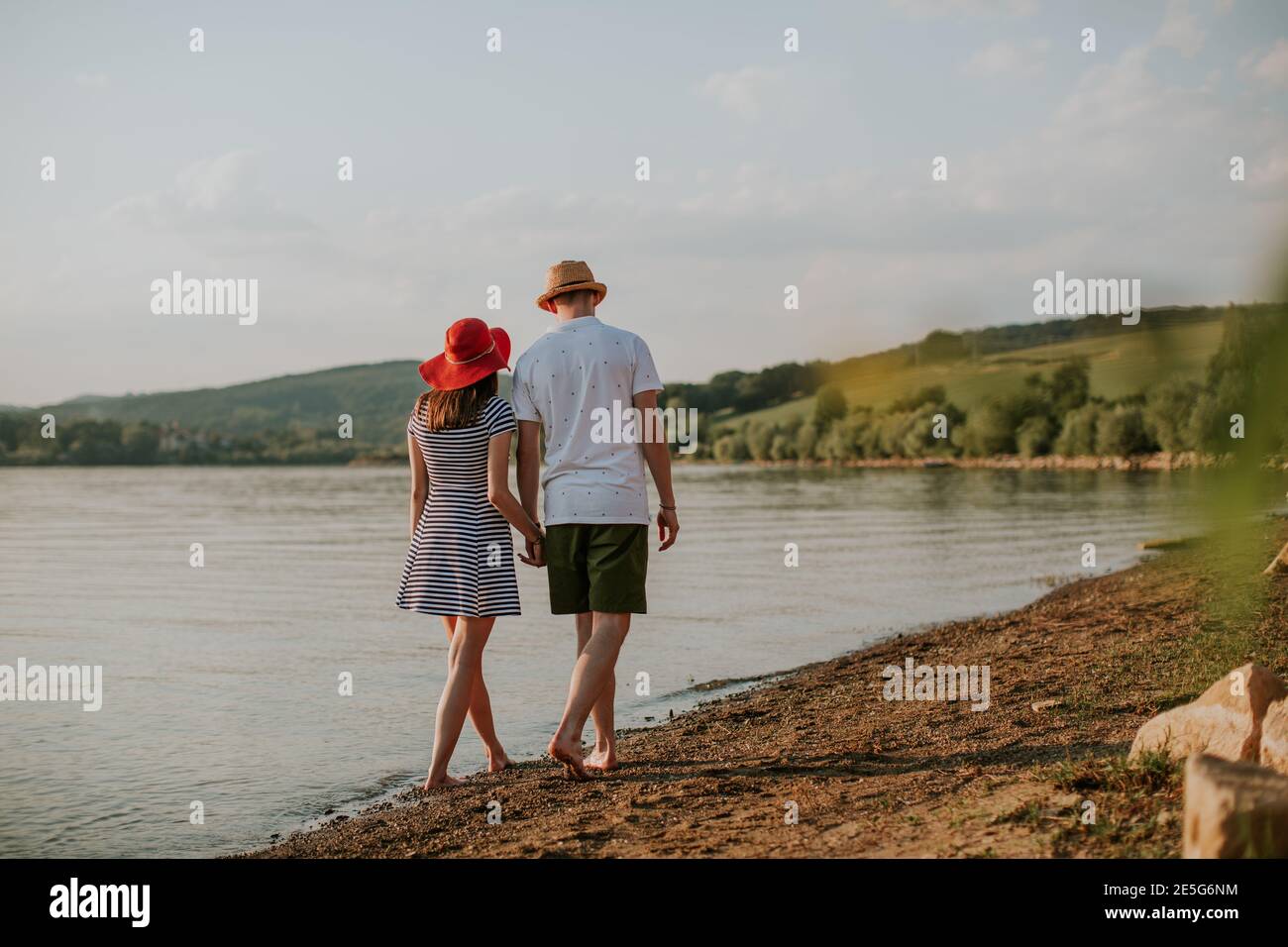 Back view of couple holding hands walking along the beach at sunset. Rear view of man and woman going for stroll at the lake on summer evening. Stock Photo