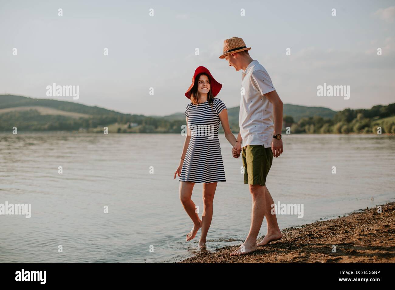 Front view of happy couple holding hands walking along the beach at sunset. Full length of smiling man and woman going for stroll at the lake on summe Stock Photo