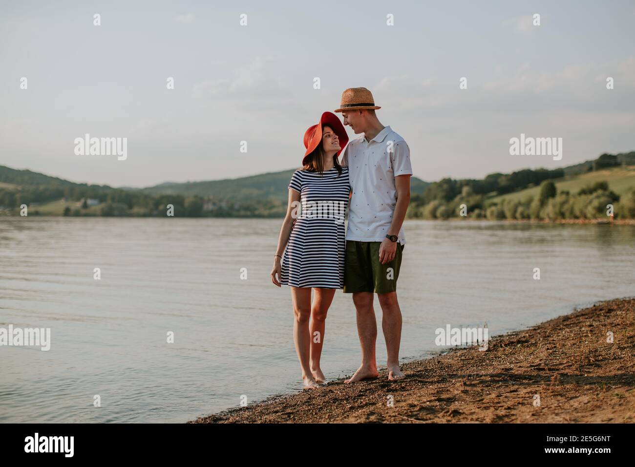 Front view of happy couple hugging while walking along the beach at sunset. Full length of smiling man and woman embracing at the lake on summer eveni Stock Photo
