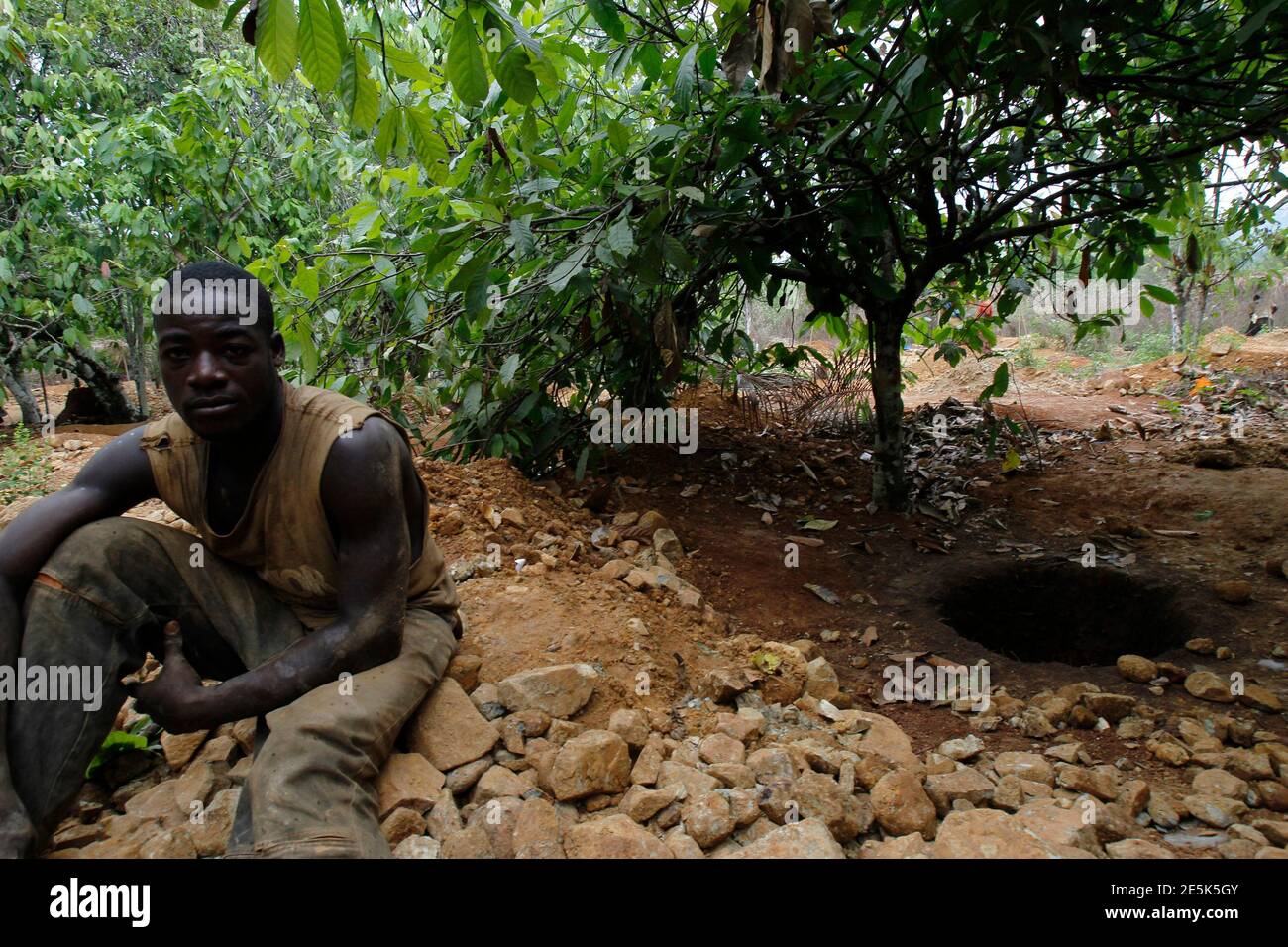 A prospector sits near a hole as he rests at a cocoa farm that is turning into a gold mine near the town of Bouafle in western Ivory Coast March 18, 2014. With high prices for the precious metal fuelling a gold rush in Ivory Coast and Ghana, diggers are scurrying to cash in. But the drain on the labour market and the harm done to cocoa plantations could endanger cocoa production in the two nations, which account for 60 percent of global supply. Picture taken March 18, 2014. To match Insight COCOA-GOLD/WESTAFRICA    REUTERS/Luc Gnago (IVORY COAST - Tags: AGRICULTURE BUSINESS COMMODITIES EMPLOYM Stock Photo