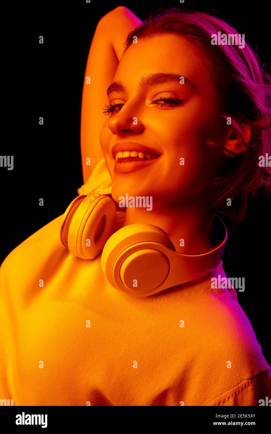 Party. Caucasian woman's portrait on black studio background in pink-orange neon light. Beautiful female model with headphones. Concept of human emotions, facial expression, sales, ad, fashion. Stock Photo