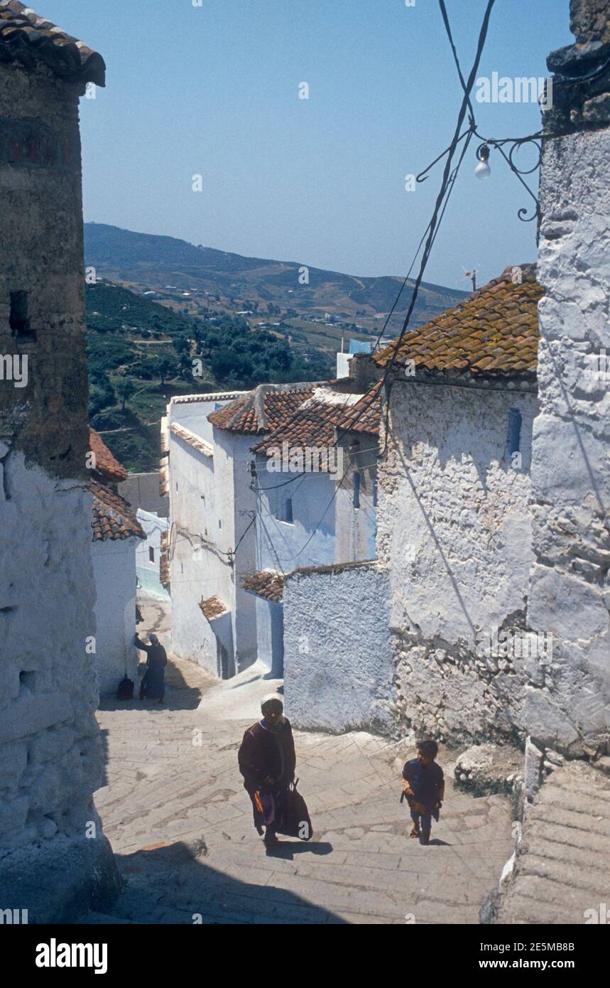 1984 Morocco Chefchaouen - Chefchaouen, Shafshāwan, also Chaouen, is a city in northwest Morocco. It is noted for its buildings in shades of blue. Chefchaouen is situated just inland from Tangier. The beauty of Chefchaouen's mountainous surroundings are enhanced by the contrast of the brightly blue painted medina (old town). People in traditional costume walking through the narrow blue painted streets of the Old town of Chefchaouen Morocco North Africa Stock Photo