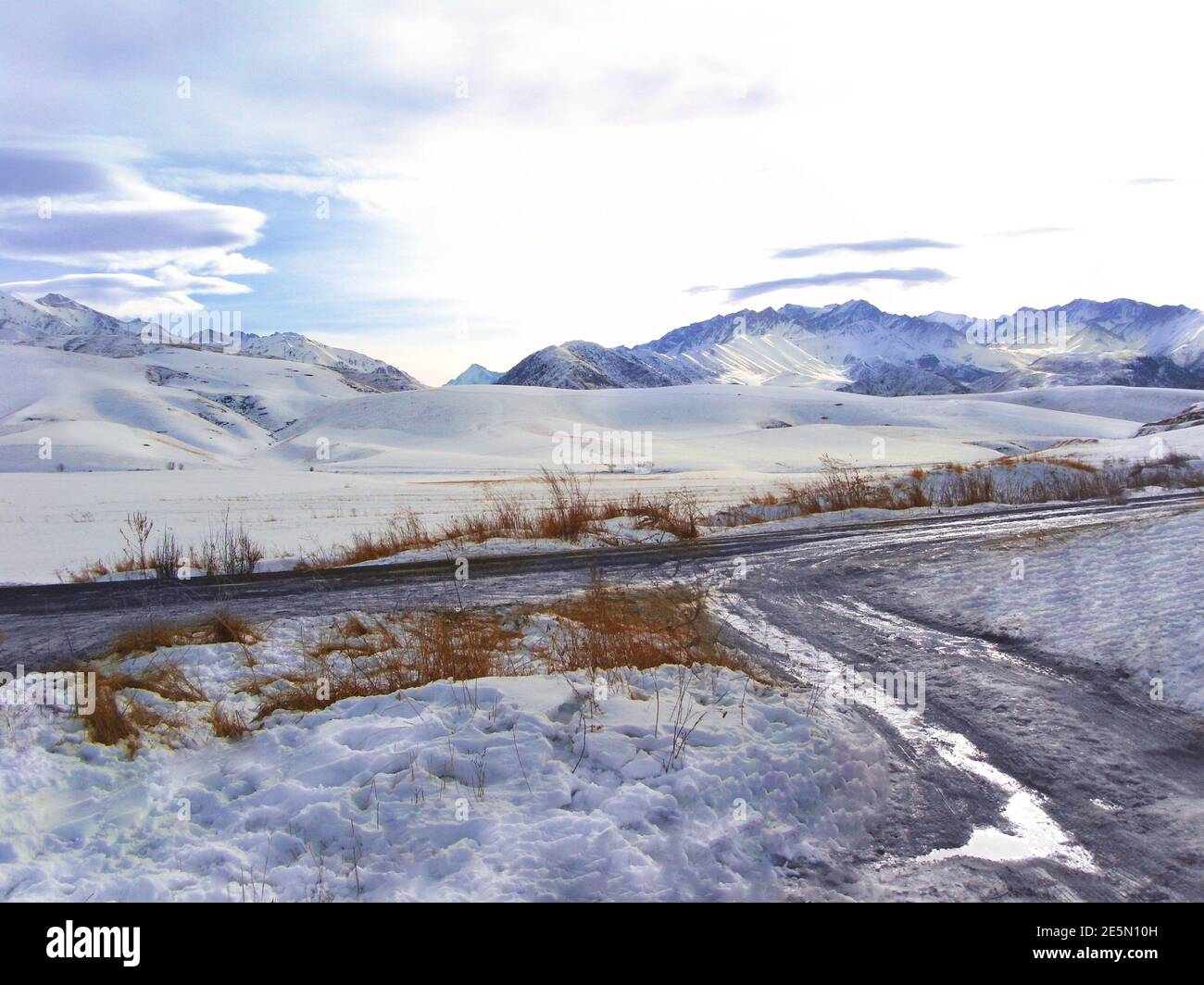 Snowy mountains and ridge, country road with puddle, brown grass in the ...