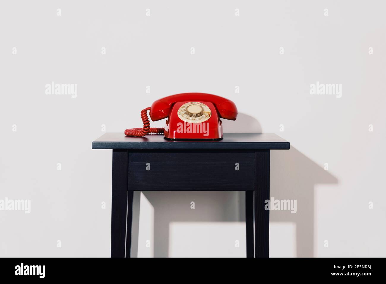 a red landline rotary dial telephone on a black wooden table, indoors, in front of a white wall Stock Photo