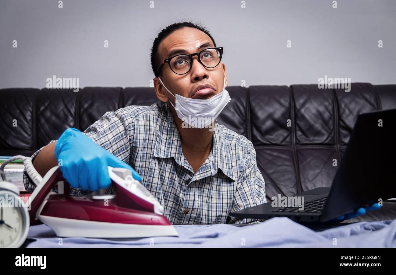 man with boring face working with very busy business and housework part, ironing cloth and working with laptop during self isolation Stock Photo