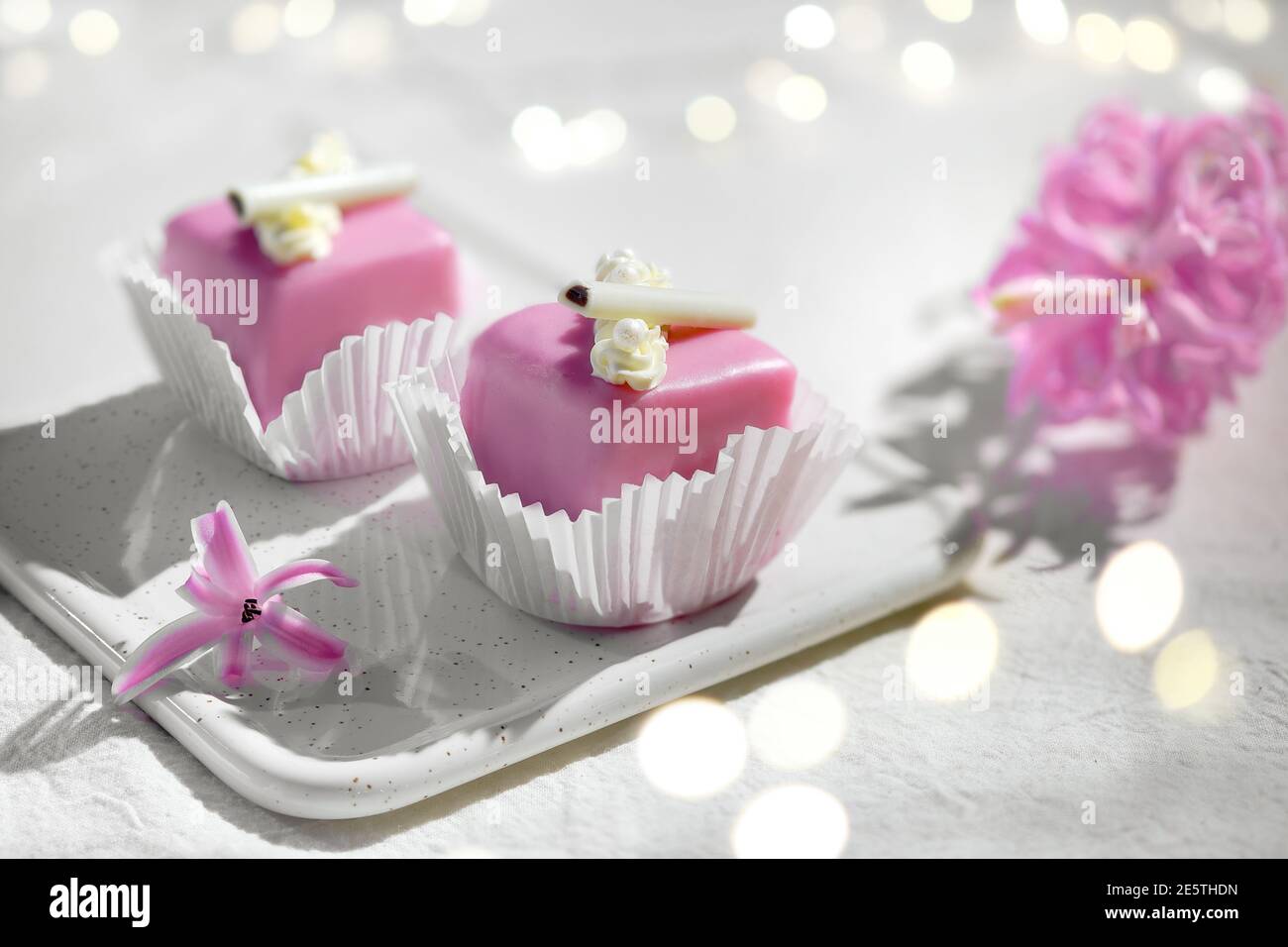 Valentine petit fours with marzipan icing. Fragrant pink hyacinth flower. Garland of lights on white textile. Happy Valentine's day Romantic evening Stock Photo
