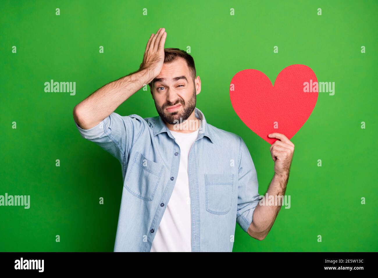 Portrait of sad guy hold red heart hand face wear blue shirt isolated on vibrant green color background Stock Photo