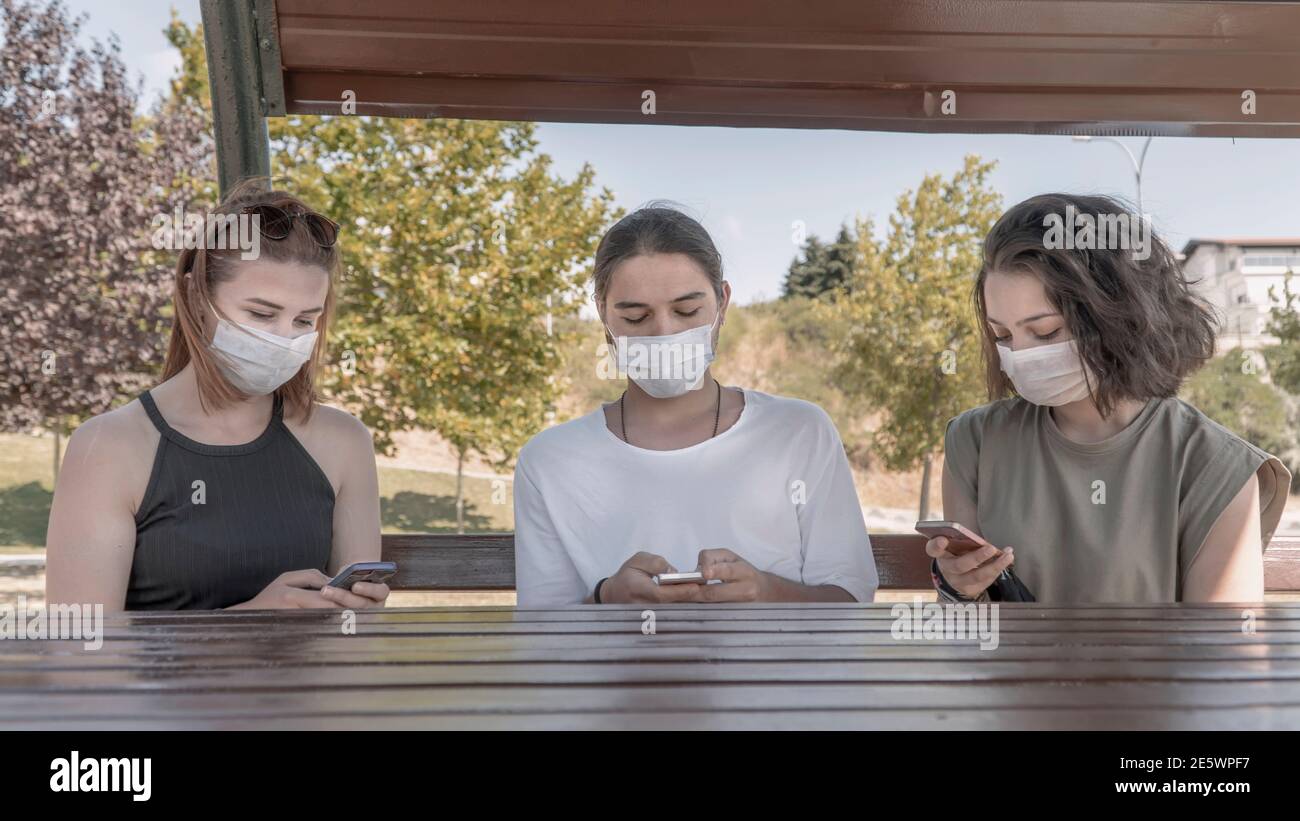 Millenials wearing face masks using mobile phone while sitting on a wooden bench in the park. Meeting with friends in the park during the coronavirus Stock Photo