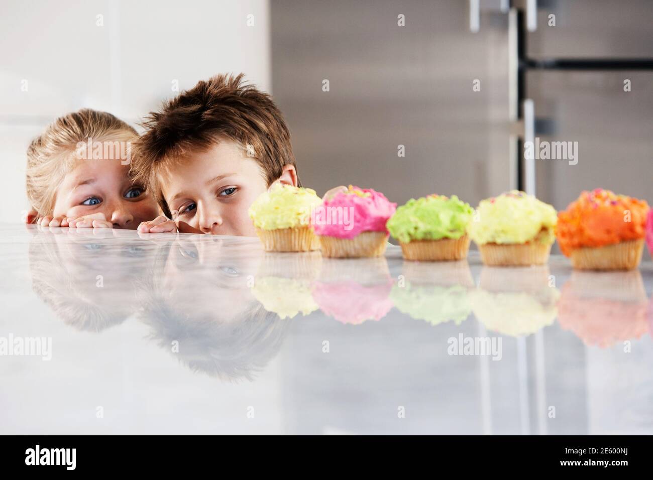 Young girl and boy peeking over counter at row of cupcakes Stock Photo