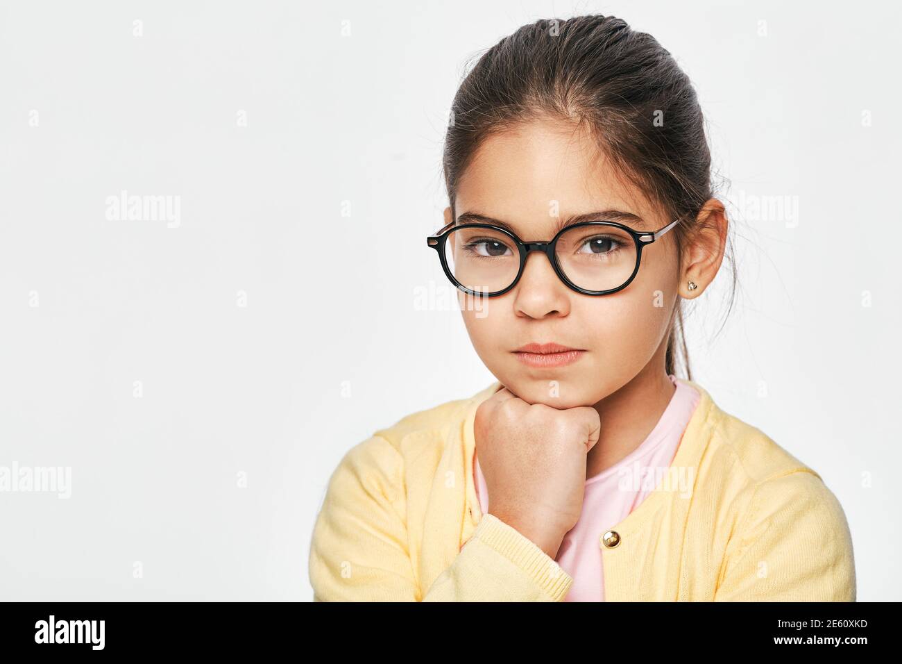 Serious Hispanic female kid wearing eyeglasses looking pensive at camera, close-up, on a white background Stock Photo