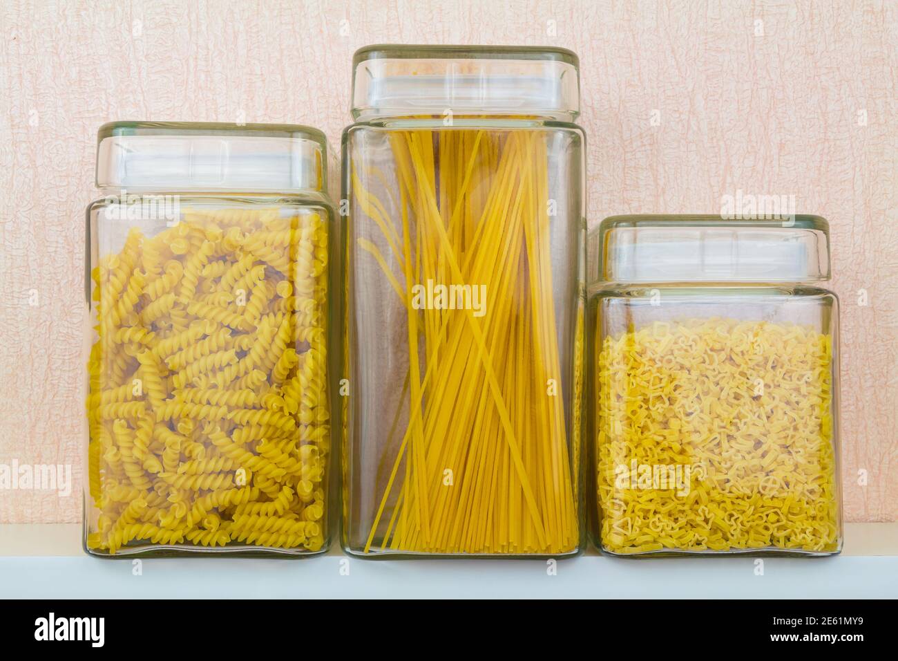 Glass jars with different shaped pasta on white kitchen shelf Stock