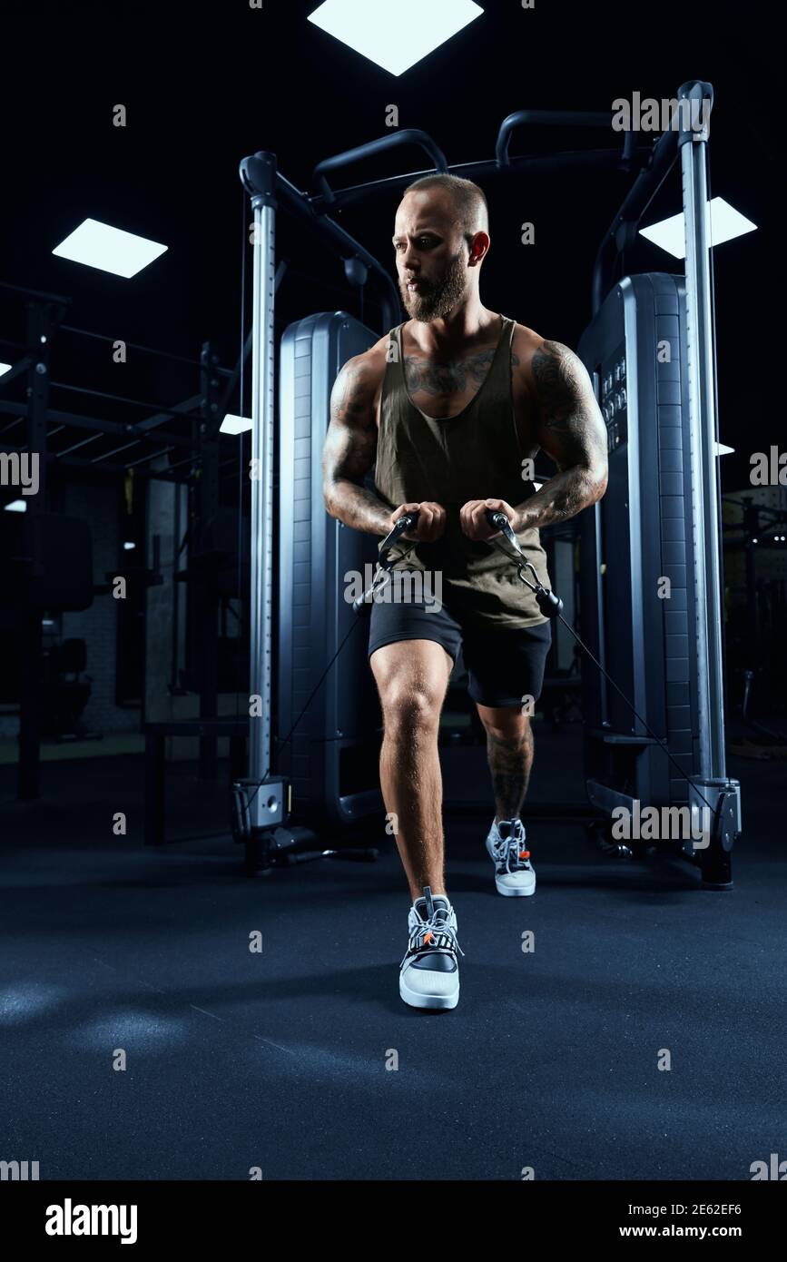 Front view of muscular bodybuilder in sportswear doing low cable crossover exercise. Portrait of man with perfect body training chest in gym in dark a Stock Photo