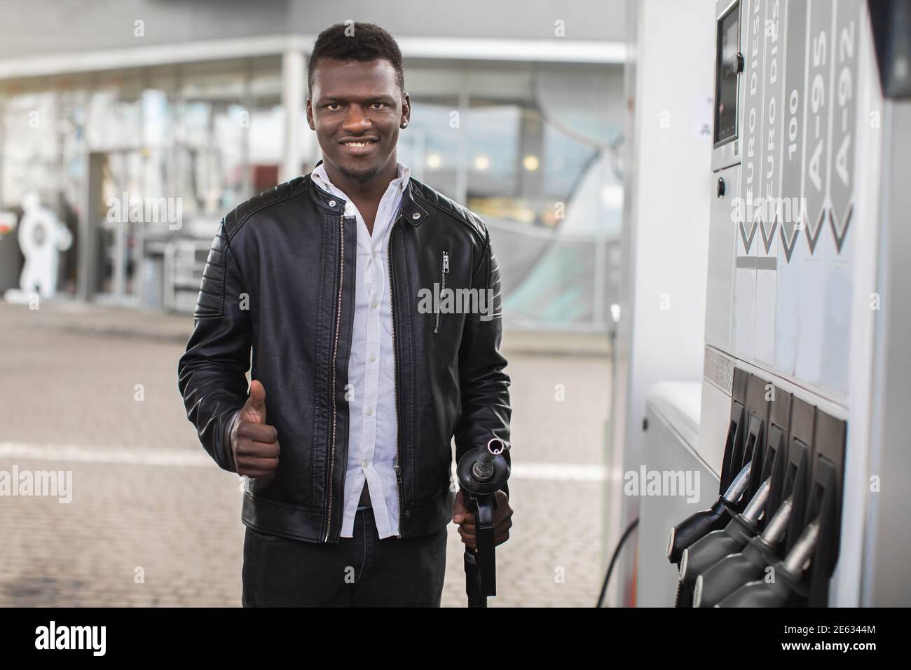 Smiling handsome young African guy, wearing black leather jacket, showing  thumb up to camera and holding filling gun at the gas station. Close up  Stock Photo - Alamy