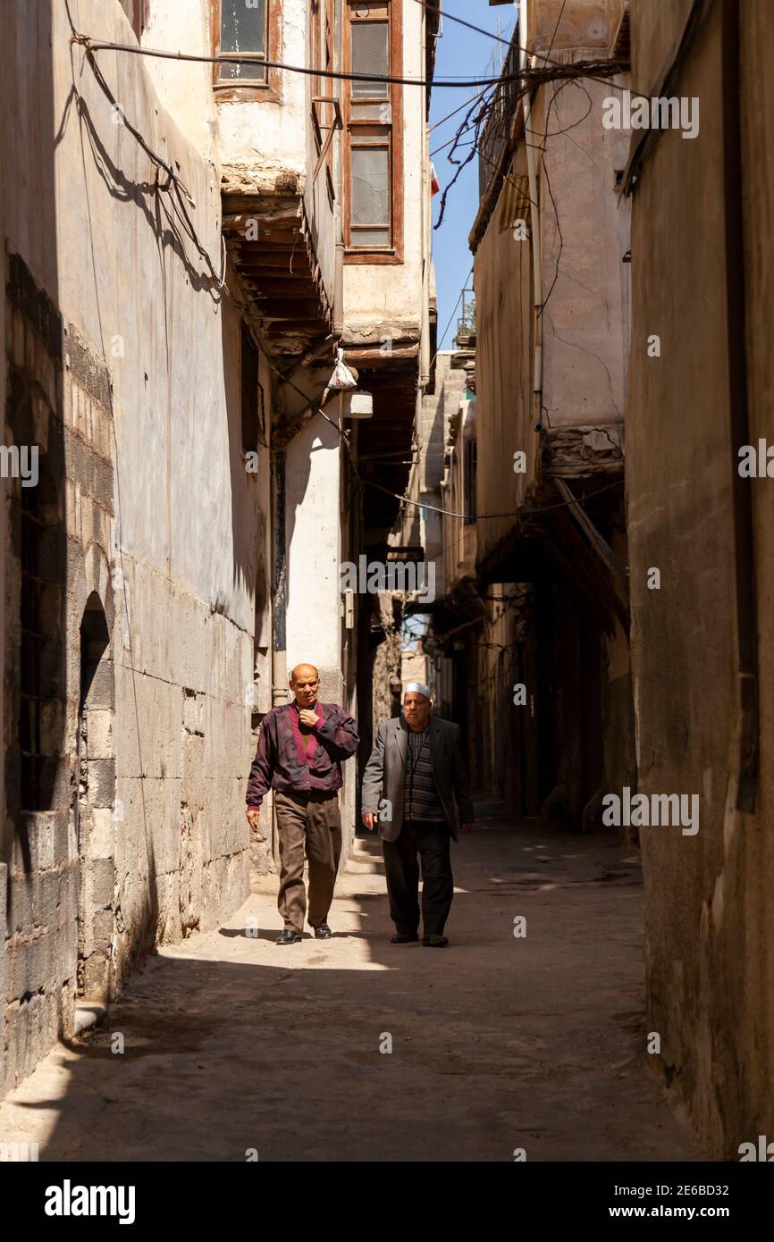 Damascus, Syria 03.27.2010: Two elderly Arabic men one wearing religious hat are walking in a narrow alley between historic houses in the old city of Stock Photo