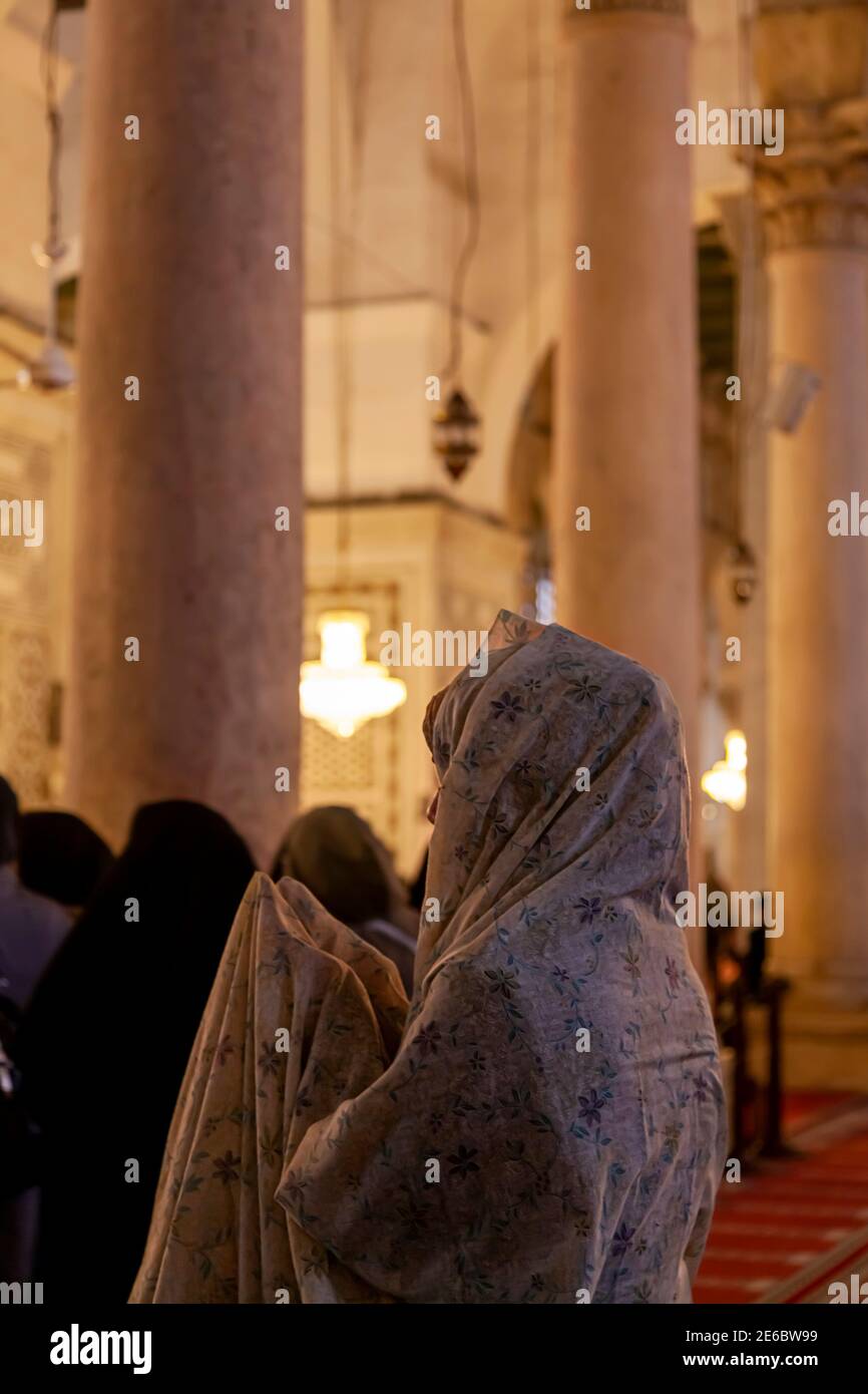 An Arabic Muslim woman wearing floral traditional head wrap is praying by raising her hands above inside a mosque in Syria. In the background there ar Stock Photo