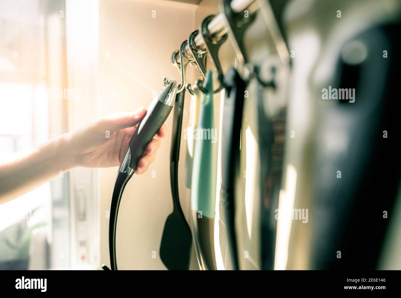 Woman hand holding black plastic handle of spatula. Woman cooking food in apartment kitchen. Cooking utensil set hanging on stainless steel wall mount Stock Photo