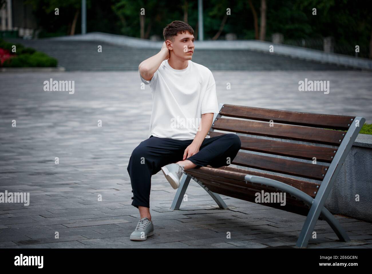 White T-shirt template on a guy sitting on a blurred background of a square, on a bench, looking into the distance, clothes front view. Mockup of men' Stock Photo