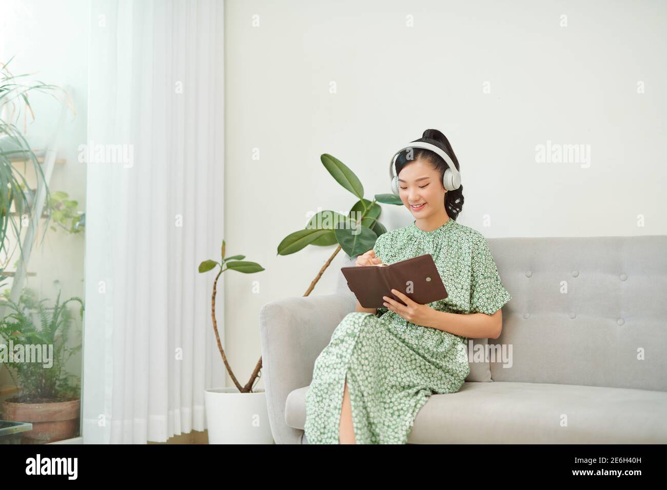Young Asian woman listening music from headphones and writing note for her work idea in diary book. Stock Photo