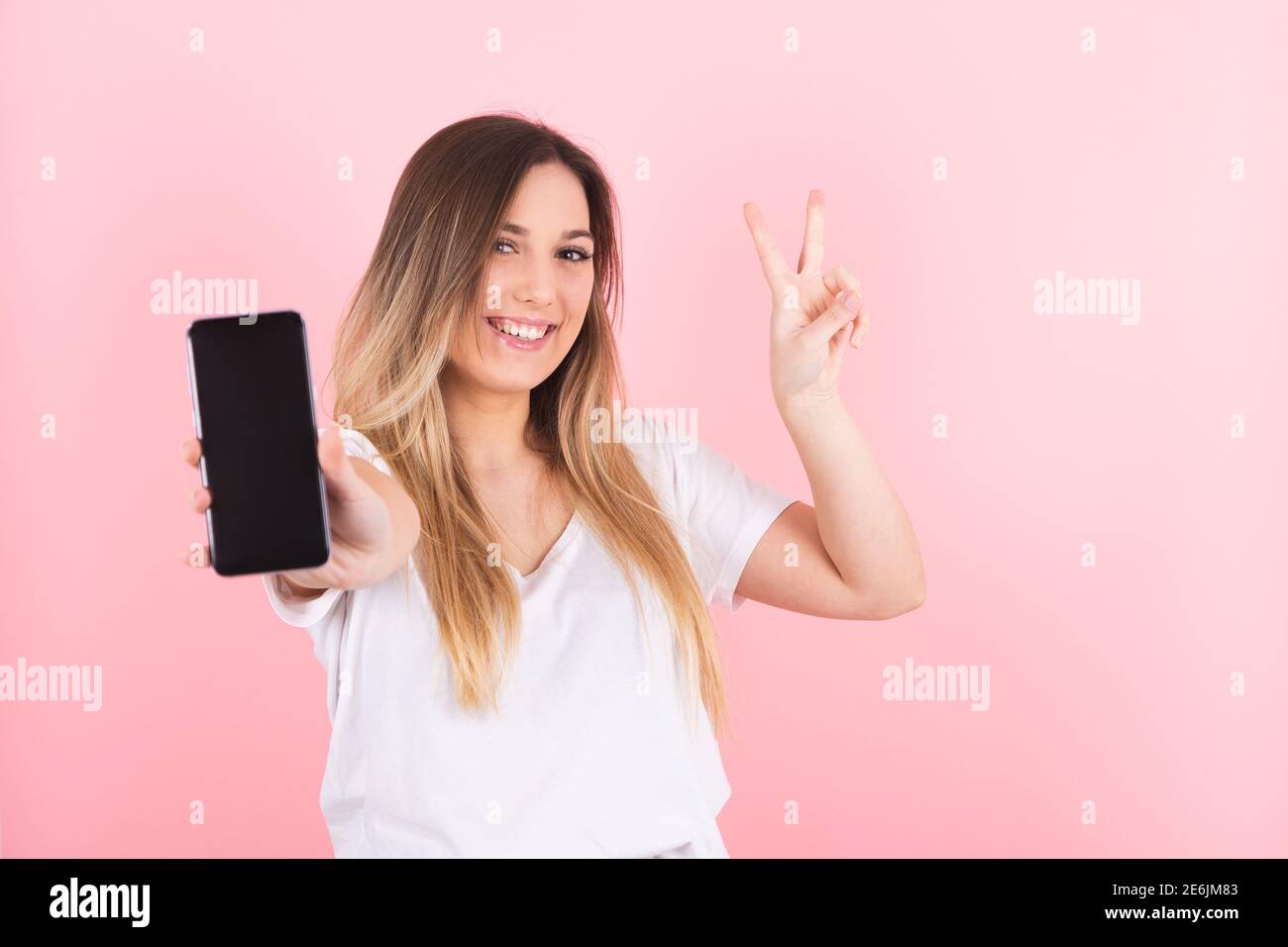a young blonde woman shows her cell phone with a victory gesture Stock Photo
