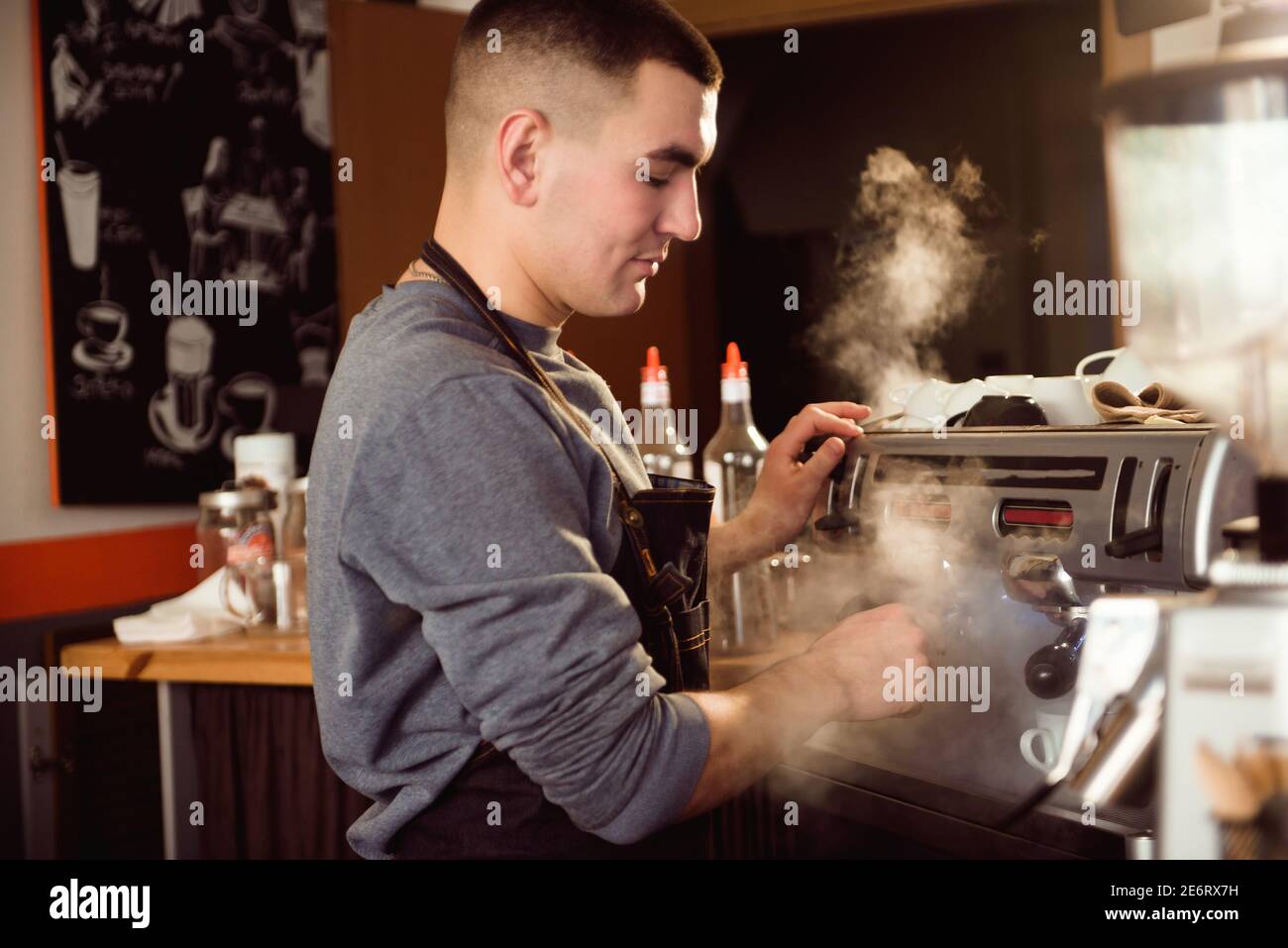 Professional barista holding metal jug warming milk using the coffee machine. Happy young man