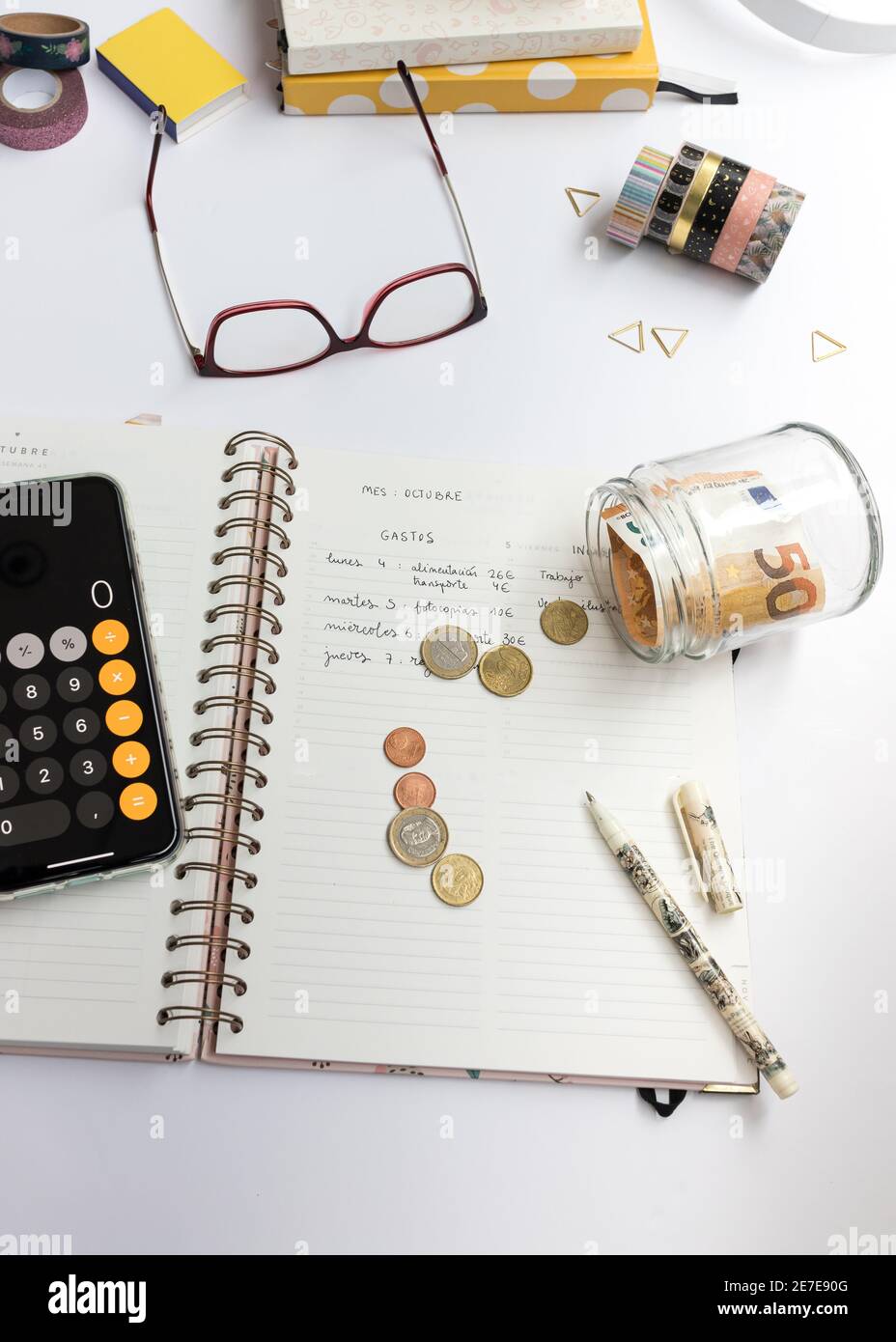 Overhead view of an opened notebook with a home budget written in it. Some coins and banknotes in a laid jar are on the notebook. A calculator is read Stock Photo