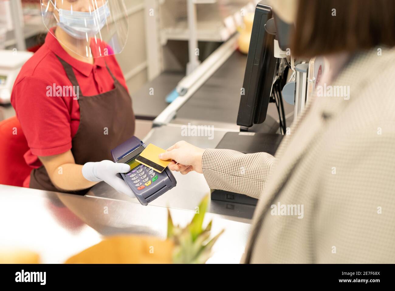 Young female cashier in protective mask, gloves and screen looking at terminal while one of consumers paying for goods by credit card Stock Photo