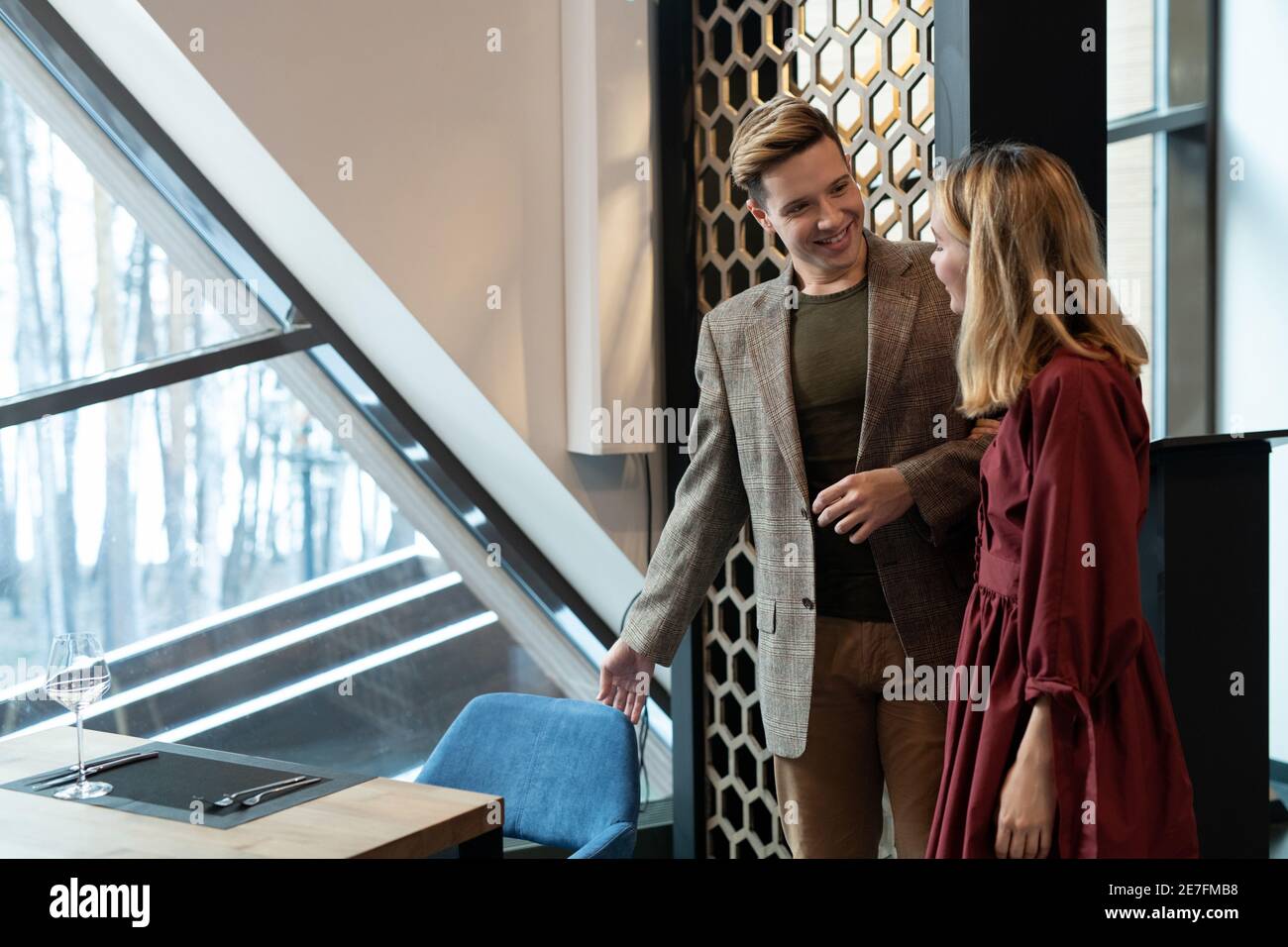Happy young elegant man in smart casualwear looking at his blond girlfriend while helping her to sit on blue velvet chair by served table Stock Photo
