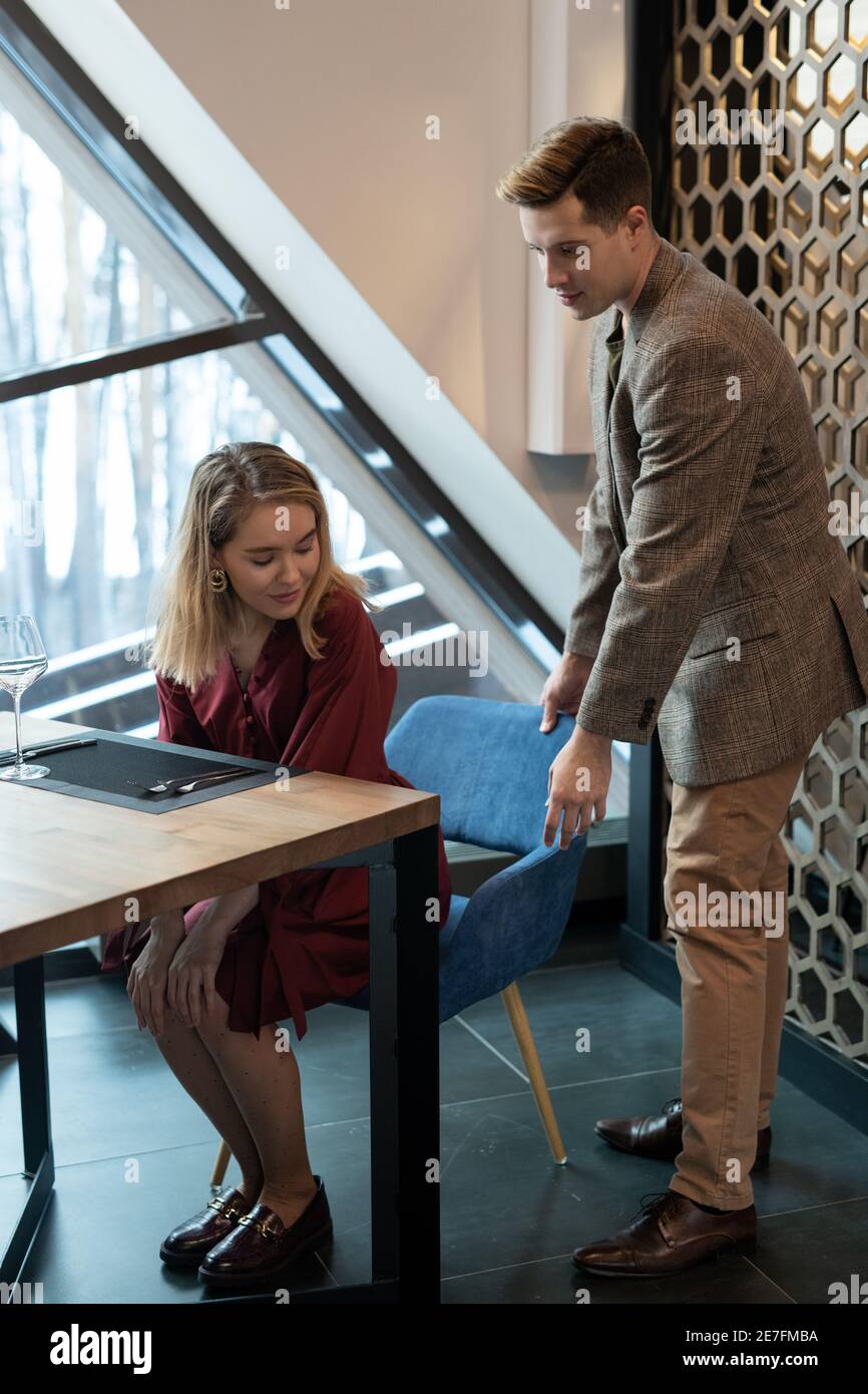 Young elegant man in smart casualwear helping his blond pretty girlfriend sit on blue velvet chair by served table in luxurious restaurant Stock Photo