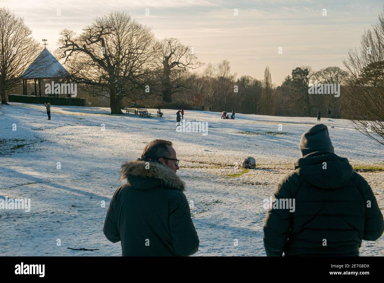 Two dads watch their children play in the snow in Golders Hill Park, London. The men wear winter clothing as the sun sets over Hampstead Heath, UK. Stock Photo