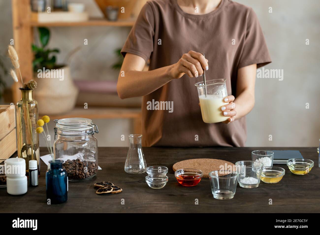 Young woman holding glassware with melted soap mass over table with glassware while making mixture for natural handmade cosmetic products Stock Photo