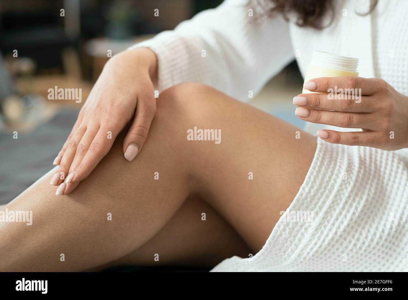 Hands of young woman in white cotton bathrobe applying natural handmade cosmetic product on her legs while sitting on bed Stock Photo