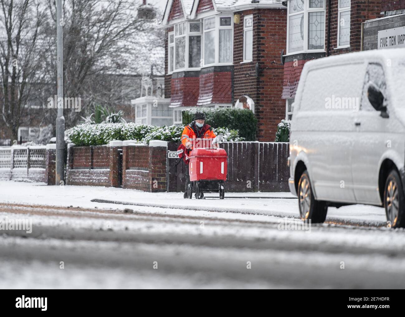 Royal Mail British post man in deep cold snow falling on road and icy frozen pavement delivering letters trolley red uniform during severe weather Stock Photo