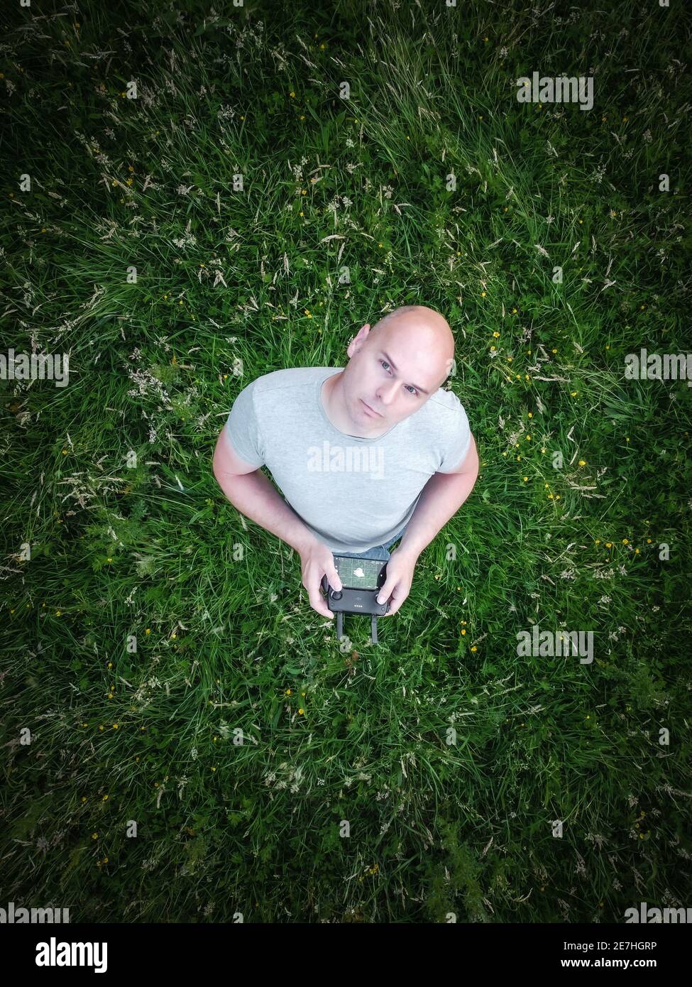 Man alone looking up at flying drone from below on ground holding controller and mobile phone in hands stood in field look to sky wearing t-shirt Stock Photo